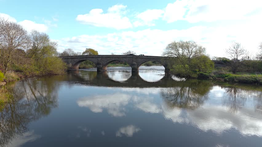 Multiple cars including compact sedan and hatchback driving along old masonry bridge above reflective water surface. Light traffic on rural crossing with natural banks and trees. Regional road