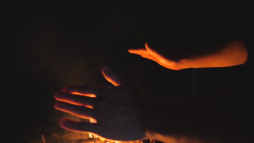 Guy warms his arms by a fire from metal brazier against the black background. Young man rubs his hands in front of an open bonfire from mangal at night. Concept of warmth and tranquil. Slow motion