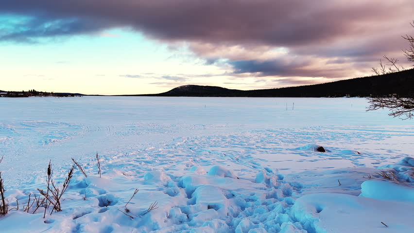 A lone snowmobile crosses a vast frozen lake in Kiruna, Sweden. Snow-covered ice fills the foreground as mountains and pine trees line the horizon under a dramatic cloudy dusk sky.