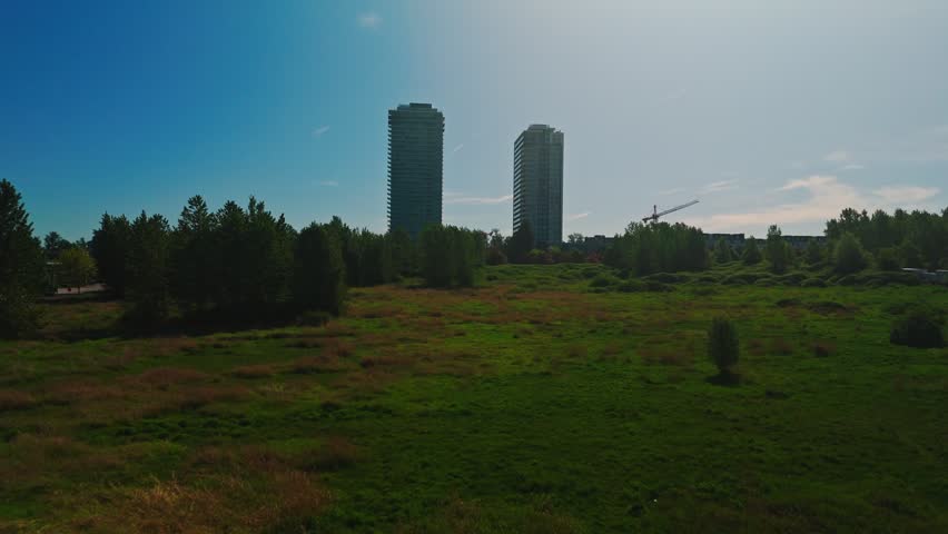Vibrant green meadow leads toward two towering concrete residential high-rises revealing yellow construction crane under clear blue sky deep afternoon shadows approaching