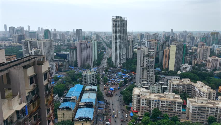 High angle aerial view of a central city highway cutting through dense residential skyscrapers and informal settlements with blue roofs in Mumbai, India.