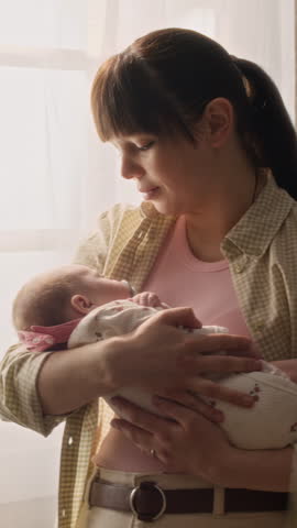 Vertical medium shot of young Caucasian mother standing by window and rocking newborn baby in her arms, enjoying moment of bonding and tenderness
