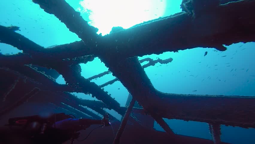 Low Angle View of Diver Levitating in Sunken Ship Skeleton at 40m. Movie-like slow motion shot from a low angle showing a scuba diver levitating inside the skeletal metal carcass of a massive shipwreck at forty meters depth. Strong light rays pierce through the surface, creating a high-contrast scene against the dark abyss. The advanced diver uses a powerful dive torch to illuminate iron frames completely covered in algae and marine growth, capturing a true deep-sea adventure.