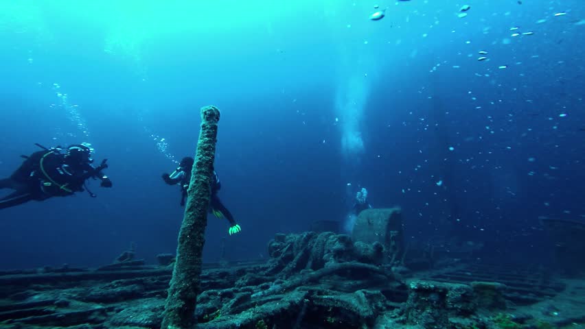 Scuba Divers Communicating by Massive Sunken Ship Wreckage at 40m Depth. Slow motion underwater pull-out shot starting close to two advanced scuba divers using hand signals near the bow of a massive shipwreck. As the camera moves away, the immense scale of the rusted vessel is revealed against the sandy ocean floor at a challenging depths from 25m to 60 meters. The dark blue, high-contrast scene features a third diver appearing, with all human figures looking tiny next to the colossal structure covered in corals, algae, and marine life amidst swirling sea currents and sediment.