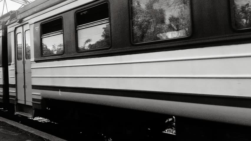 Black and white view of a passenger train moving along railway tracks in an outdoor setting. The composition captures motion and perspective, with the train extending into the distance and overhead electric lines framing the scene. Reflections in the windows and blurred surroundings enhance the sense of movement and atmosphere. The monochrome style adds a cinematic and timeless mood. The scene represents travel, transportation, and urban infrastructure. Ideal for themes of trains, journeys, motion, and classic aesthetics.