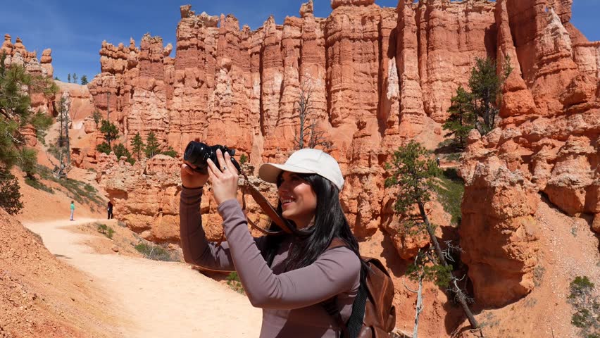 Happy female photographer with a backpack taking pictures of the stunning red rock formations while hiking on a trail in bryce canyon national park, utah, during a sunny summer day