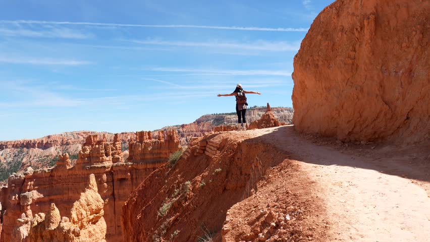 Young female traveler with a backpack enjoying the stunning panoramic view of bryce canyon, raising her arms in a moment of freedom and achievement while standing on the edge of a scenic trail
