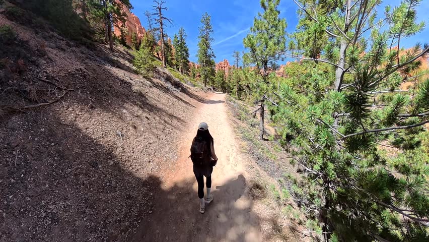 Young female traveler hiking down a dusty trail in bryce canyon national park, utah, surrounded by red rock formations, pine trees, and a clear blue sky on a sunny summer day