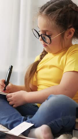 A girl wearing glasses sits on the floor, writing intently. The girl is focused on her work with a notepad next to her in a bright, sunlit room.