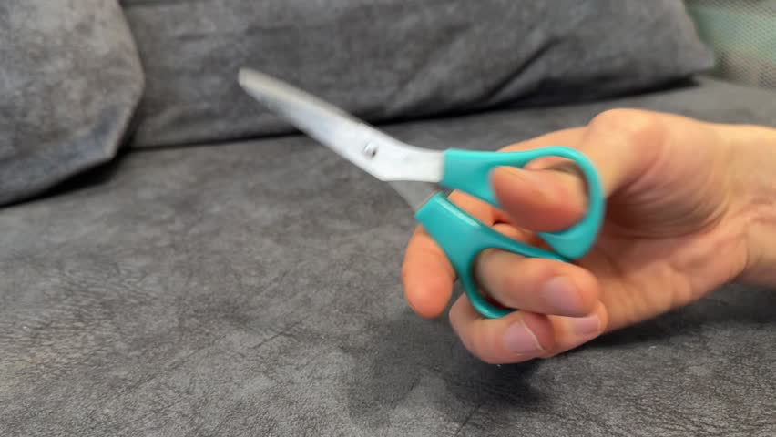 Hand holding small scissors while sitting on sofa indoors. DIY concept
