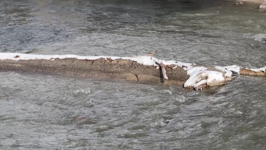 Flowing river water near a narrow concrete barrier with small patches of snow outdoors, winter nature scene, quiet landscape, cold season.