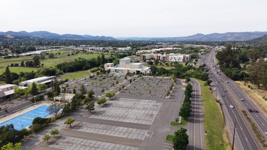 Panning aerial view of Napa Valley College campus with empty parking lot, outdoor swimming pool and adjacent highway with moving traffic against green hillside backdrop