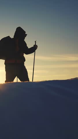 A man stands silhouetted against a sunset, holding a pole. The man is prepared for winter hiking in a snowy landscape with soft white snow below.
