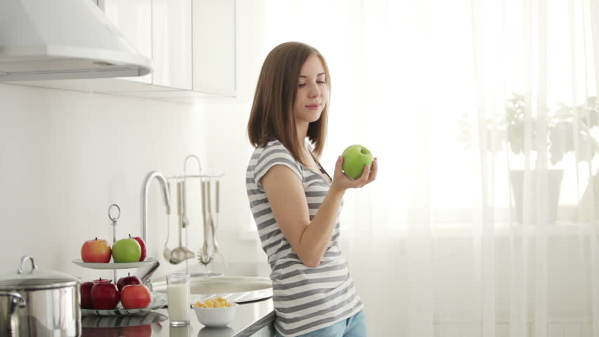 Cute girl standing in the kitchen and eating an apple

