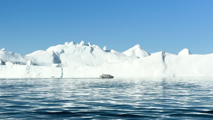 A boat moves through water close to a large glacier. Icy mountains rise in the background. The sun shines. The view shows a winter landscape with clear skies.