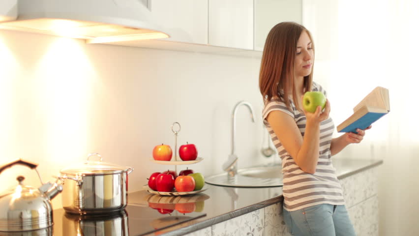Cute girl standing in kitchen reading book and eating apple
