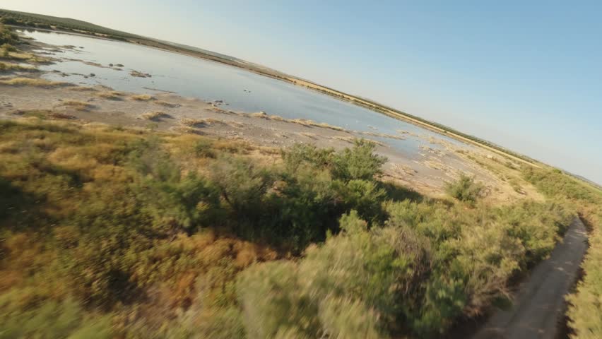 Expansive view of a shallow lake and vast marshland under a bright, clear blue sky. Dry vegetation and small bushes line the muddy banks. Several birds move across the calm water.
