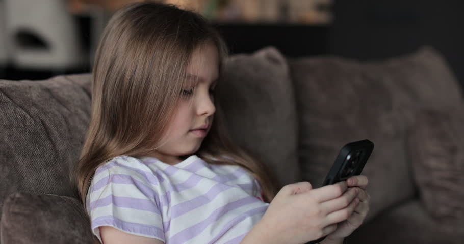 Female Caucasian teenager kid using mobile phone at home in living room sits on sofa. The problem of addiction to gadgets.