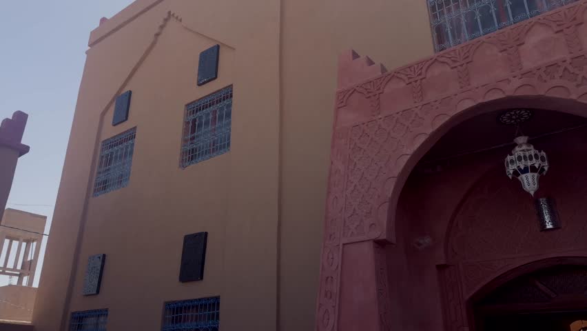 Carved red plaster mosque arch with decorative lantern beside a beige building with barred windows in Morocco