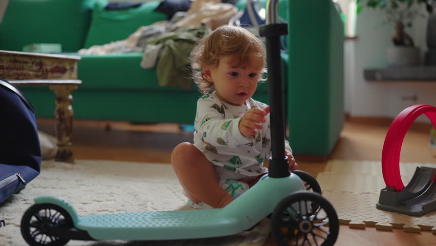 Young child on floor holding scooter and looking up with attentive expression, representing observation, engagement, discovery and relaxed family home environment