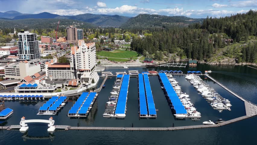 Coeur d Alene, Idaho resort and marina with blue boat slips on lake with downtown buildings and lush green pine trees under bright blue sky in Pacific Northwest United States. Aerial orbit wide shot.