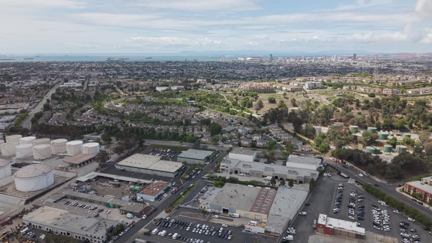 Long Beach industrial area with oil storage tanks, warehouses, residential neighborhoods, freeway and distant downtown skyline with harbor under hazy overcast sky. California, USA. Aerial wide shot.