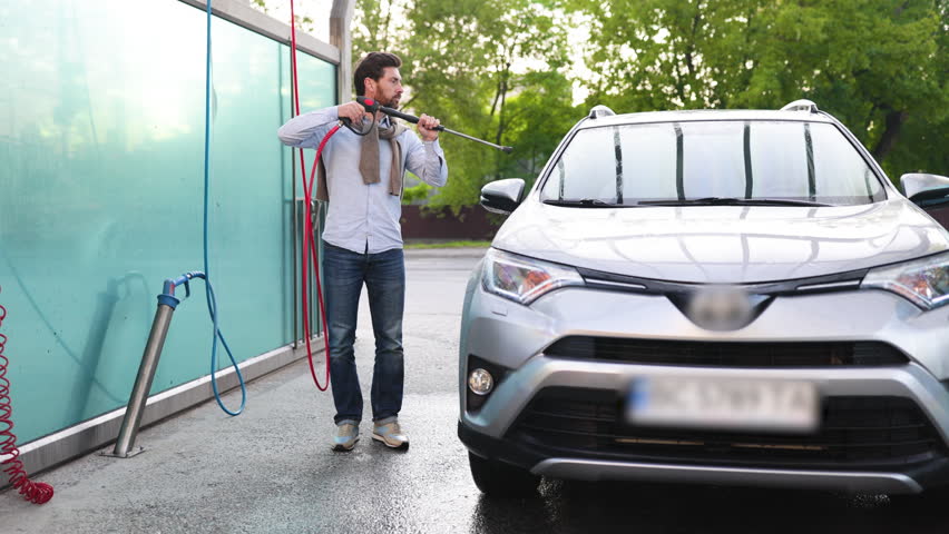 A man is actively cleaning his silver SUV at a self-service car wash station using a high-pressure water spray