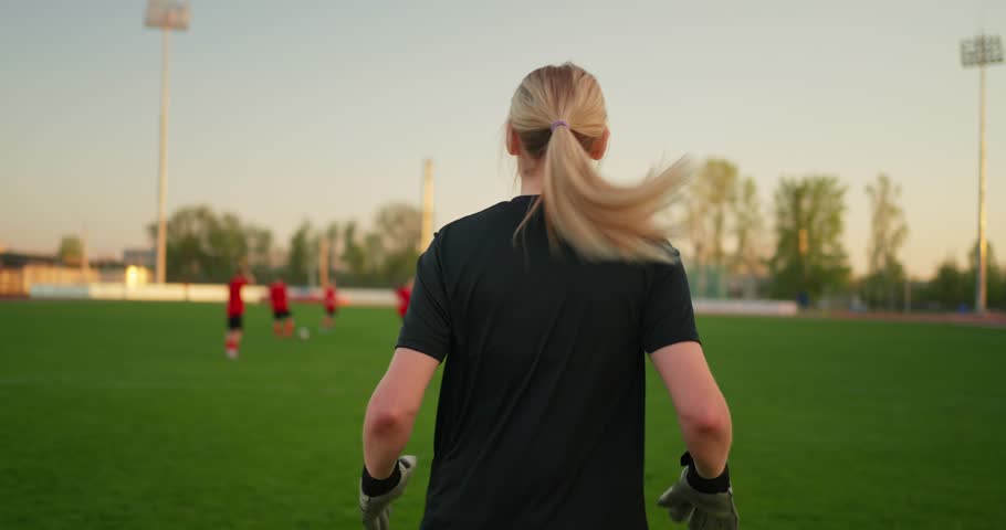 Blonde female soccer goalkeeper in black training kit and gloves on a green field during outdoor practice with teammates in the distance