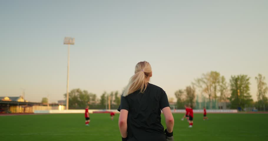Blonde female soccer player seen from behind watching teammates train on a green field during warm evening practice under an open sky