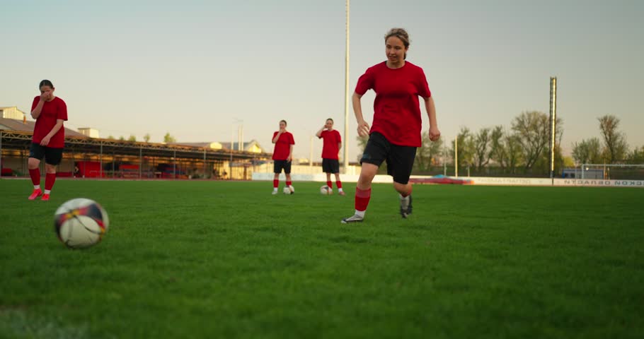 Youth soccer players in red uniforms training on a green outdoor field at sunset, running drills and chasing a ball during team practice