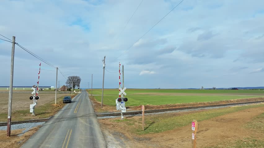 Cars wait at a train crossing where lights flash and barriers lower. The setting is a rural area with open fields in sight. The weather appears cloudy and cool.