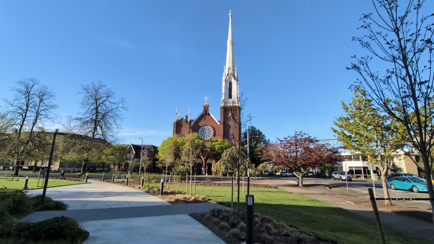 Historic brown church with a soaring white steeple, glass façade, park greenery, and passing traffic under a blue sky, showcasing community life and architectural beauty.