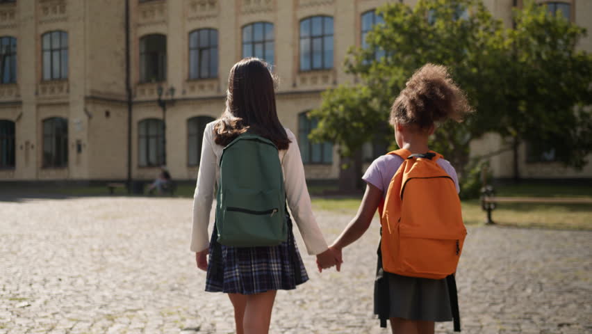 Back view two girls schoolgirls with backpacks go to school together friends females African American children little kids elementary class education studying classmates students pupils walk outdoor