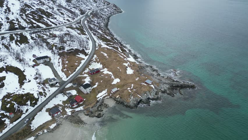 Aerial view of a winding coastal road and traditional houses along the snowy shores of Grotfjorden, Troms, Norway.