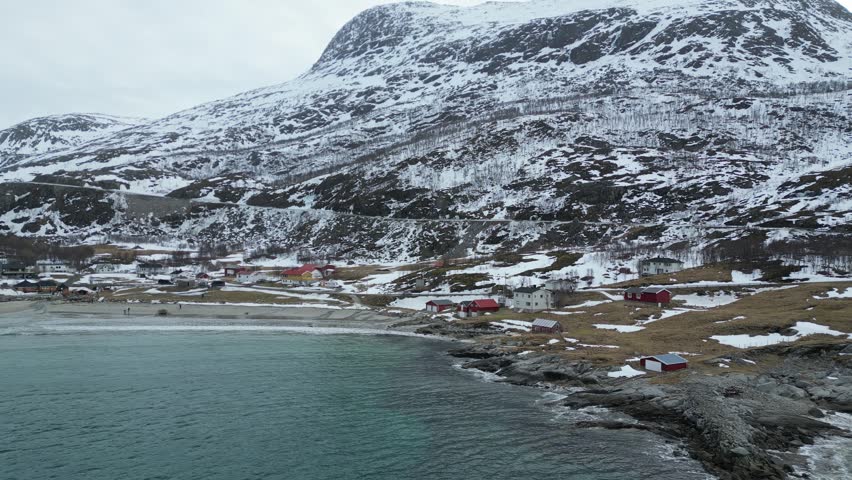 Aerial view of the coastal village of Grotfjorden with red houses and snow-covered mountains under a cloudy sky Grotfjorden, Troms, Norway.