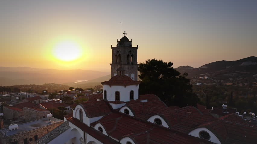 Traditional Cypriot village church bell tower silhouette against a vibrant sunset sky, rolling mountains and houses in the elevated landscape of Lefkara. Travel to Cyprus. Aerial view drone flight