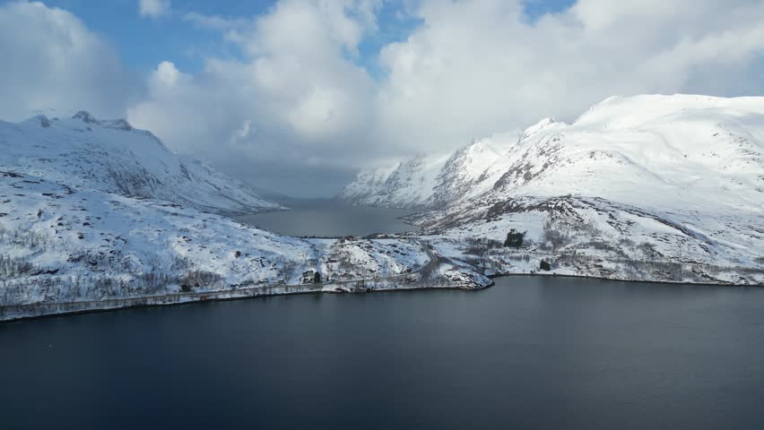 Aerial view of the Grotfjorden fjord surrounded by snow-capped mountains and a winding coastal road under a cloudy sky in Grotfjorden, Troms, Norway.
