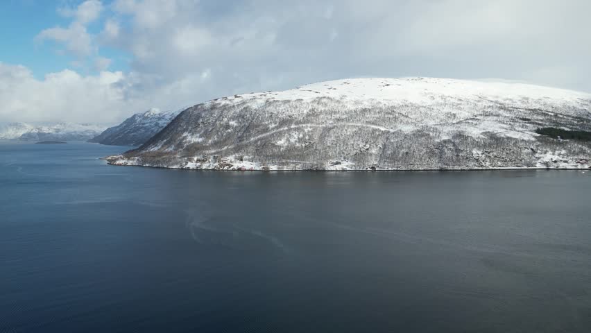 Aerial view of Grotfjorden fjord with snow-covered mountains and deep blue water under a cloudy sky in Grotfjorden, Troms, Norway.