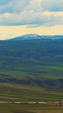 Vertical landscape shot of green agricultural fields and distant hills under cloudy sky.