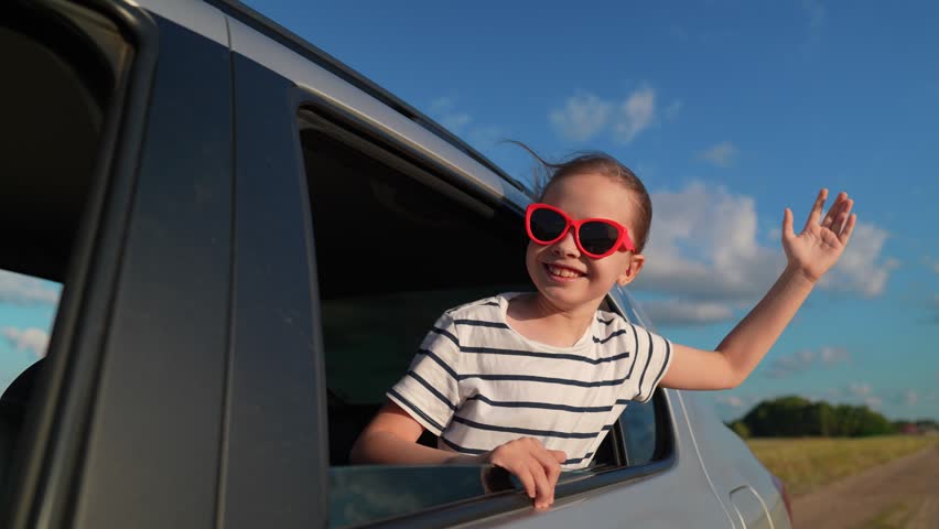Smiling child girl wearing sunglasses travels in family car. Happy child girl waves car window. Summer car vacation concept family trip with children. Childhood dreams, vacations kid. Family car trip
