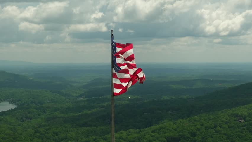 Flag waves on a mountain top with green valleys and clouds in the background in the United States during daytime