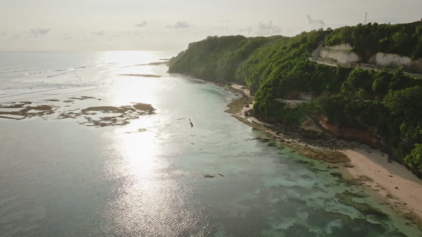 Aerial view Green Bowl Beach in Bali, Indonesia, tropical coastline with lush green cliffs, clear turquoise ocean water, and sunset reflections shimmering over the shallow reefs. Drone flight footage