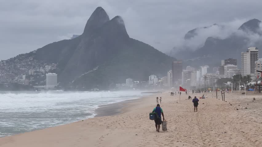Aerial view of Ipanema Beach with people walking on the sand near crashing waves and misty mountains under a cloudy sky in State of Rio de Janeiro, Brazil.