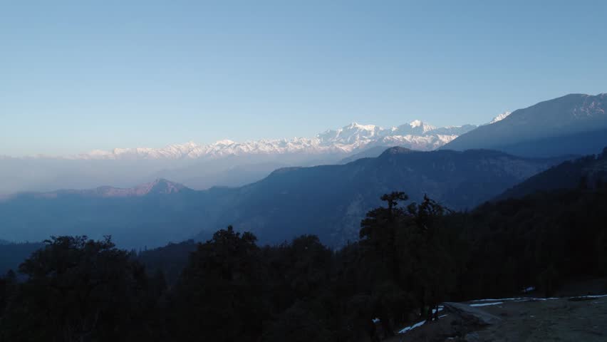 Aerial view of the snow-capped Himalayan peaks of Chopta Tungnath above dark evergreen forests under a clear blue sky in Chopta, Uttarakhand, India.