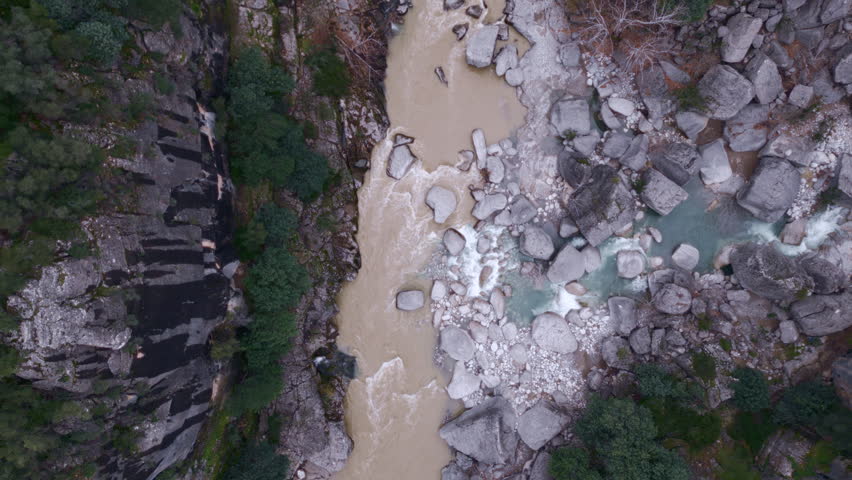 Top down drone view of a muddy river running through a rocky canyon with green vegetation.