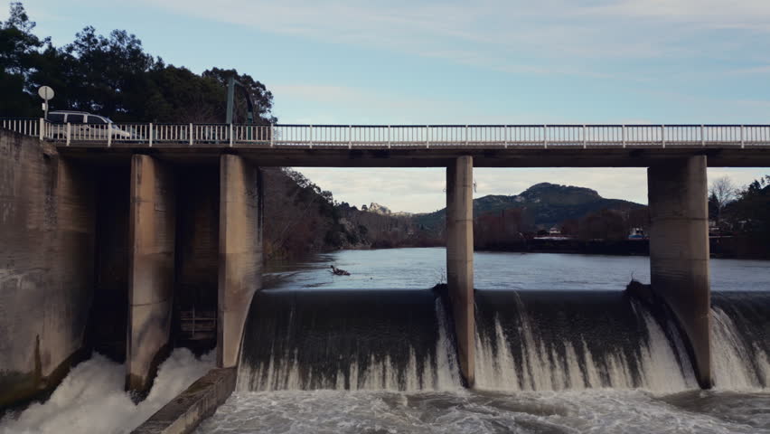 Bridge and dam over flowing water captured from a low aerial angle in a natural outdoor setting.