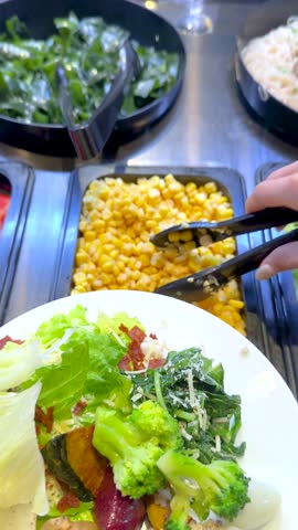 A hand uses black tongs to add sweet corn to a healthy vegetable salad plate