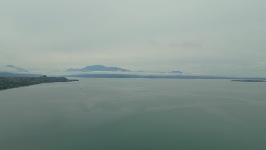 Wide aerial view of calm lake surface with distant mountains and cloudy sky