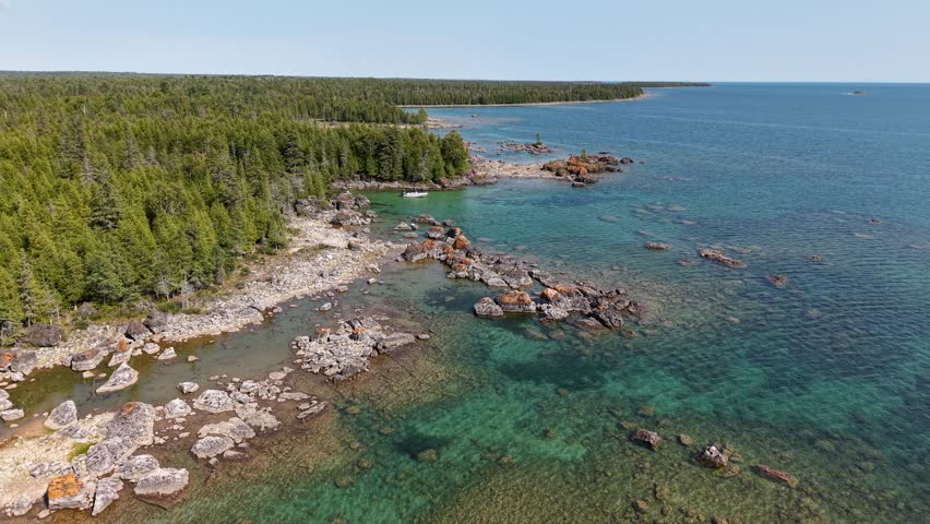 Drone aerial footage of a rocky shoreline and clear waters near Hessel, Michigan, with the Les Cheneaux Islands in Lake Huron visible in the background under bright daylight conditions.