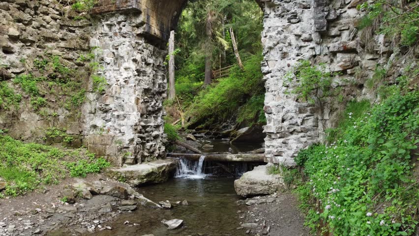 Small mountain stream flowing under the stone abandoned half-destroyed narrow-gauge railroad bridge in a forest, aerial view while moving forward on a low altitude in overcast day
